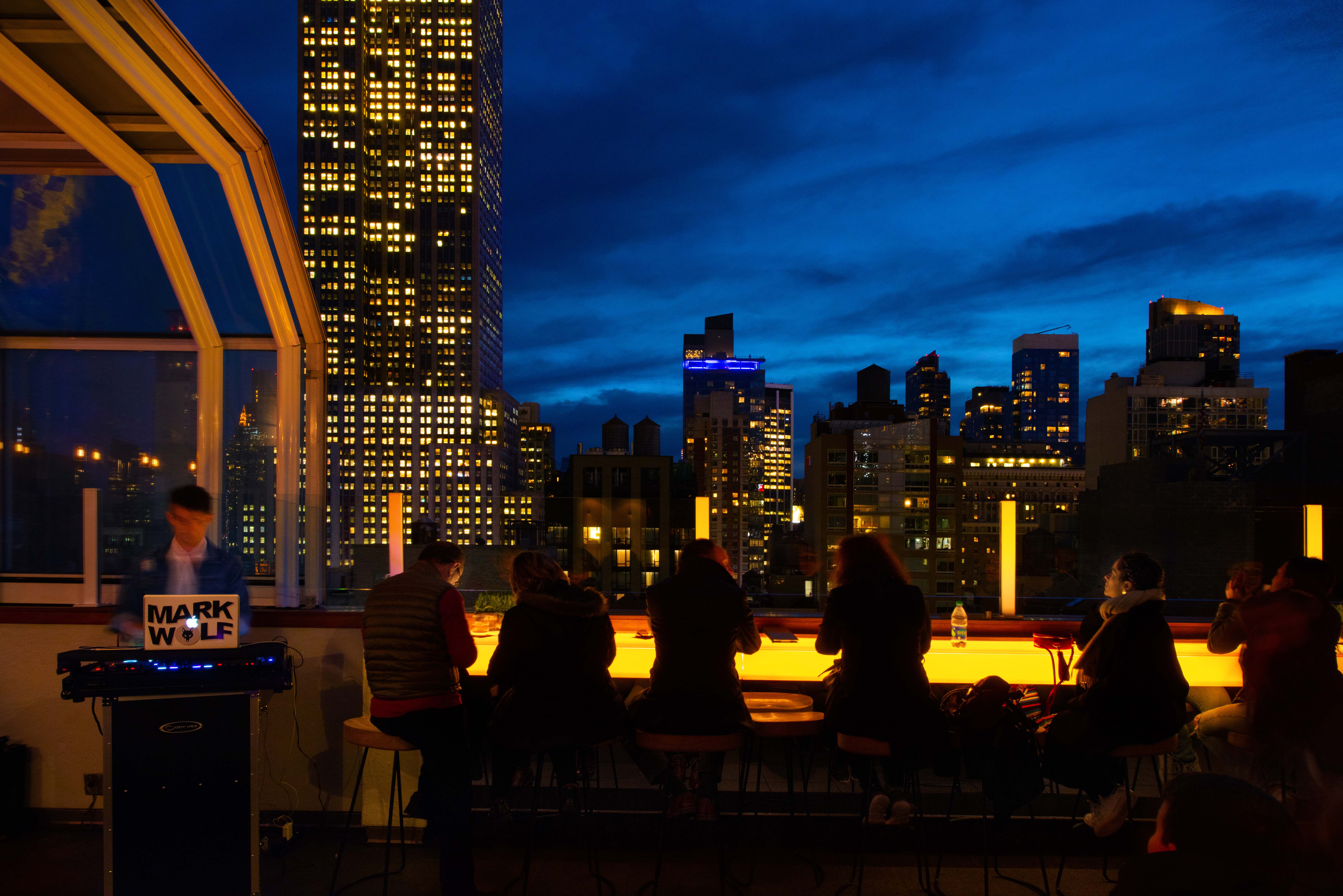 People enjoying rooftop bar with view of Manhattan at night as DJ plays music